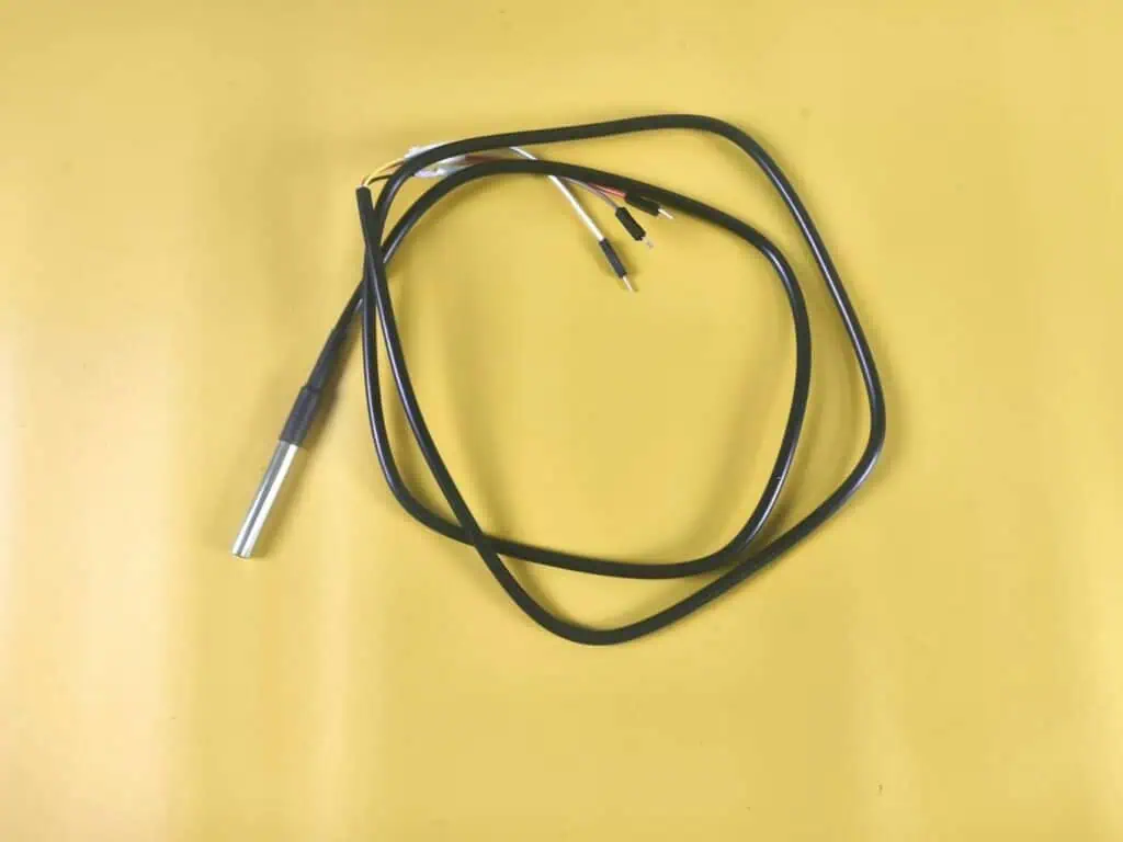 A waterproof DS18B20 temperature sensor featuring a stainless steel probe tip and coiled black cable with three connector pins (VCC, GND, Data) for liquid IoT monitoring.