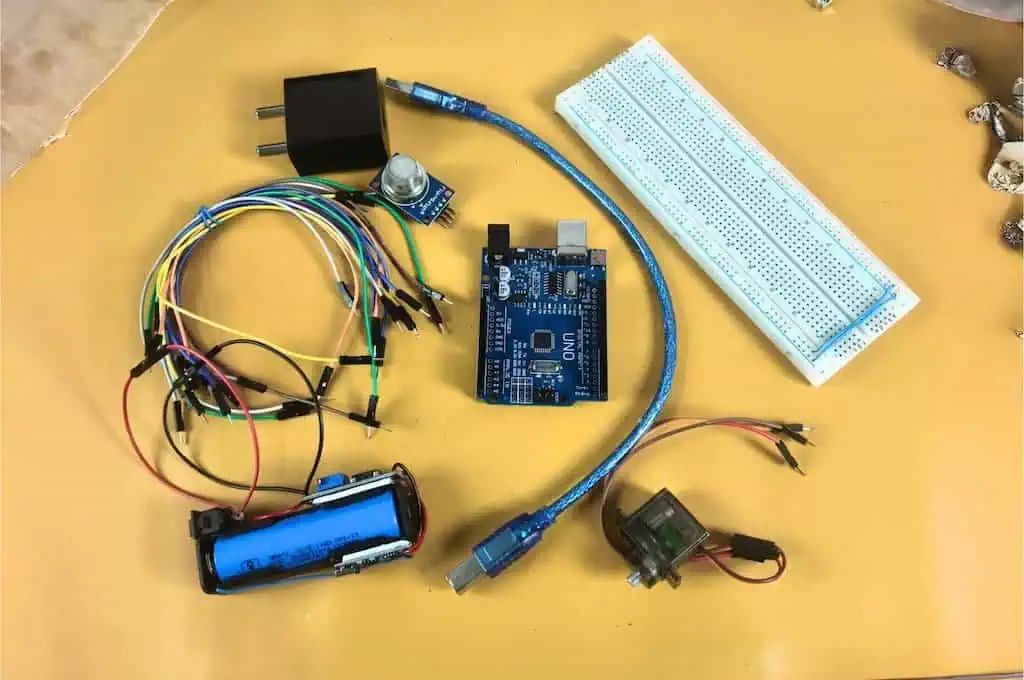 Flat lay of electronic components for the LPG gas leakage detector project, including Arduino Uno, breadboard, MQ-2 gas sensor, MG90S servo motor, and jumper wires on a yellow table.