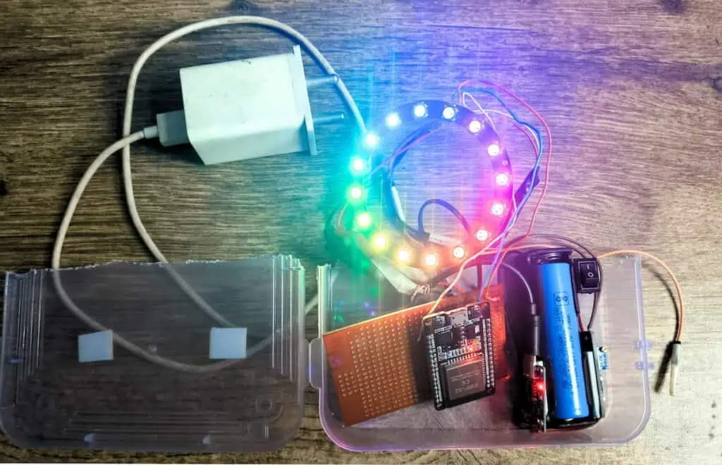 Devraj's actual ESP32 LED ring build — WS2812 16-LED ring glowing in rainbow colours, mounted with an ESP32 board and perfboard inside a clear plastic enclosure, powered by a USB phone charger, with a blue 18650 battery and rocker switch visible.